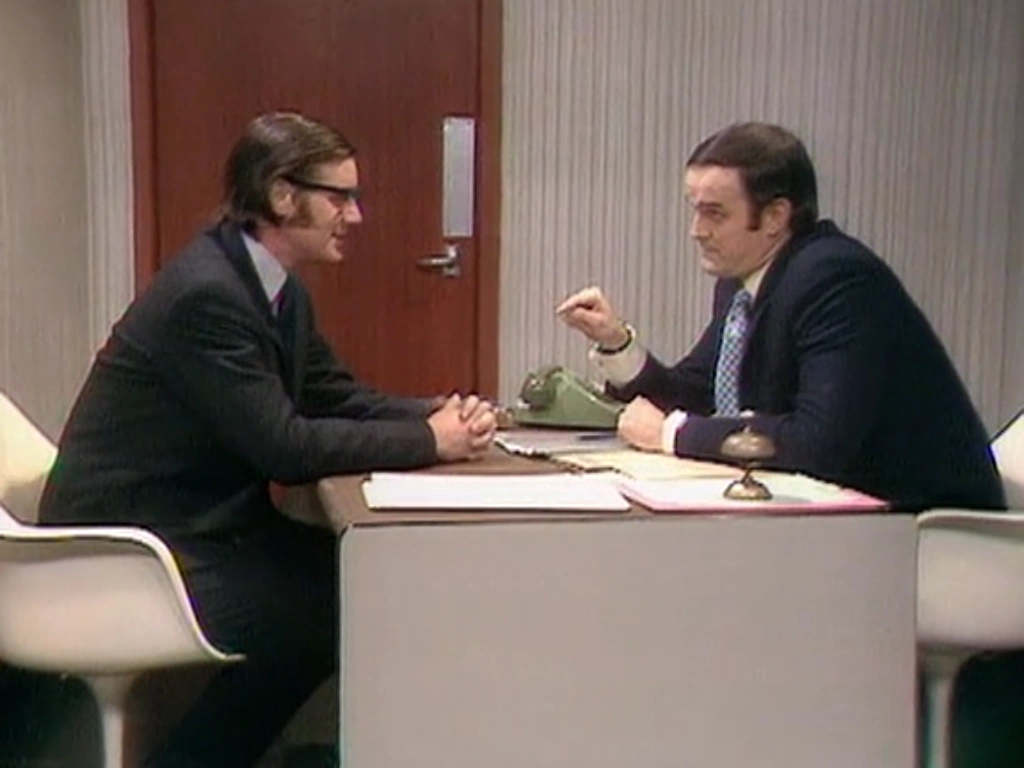 Still from the Monty Python sketch "Argument Clinic" featuring Michael Palin and John Cleese having an argument while seated across from each other at a desk.