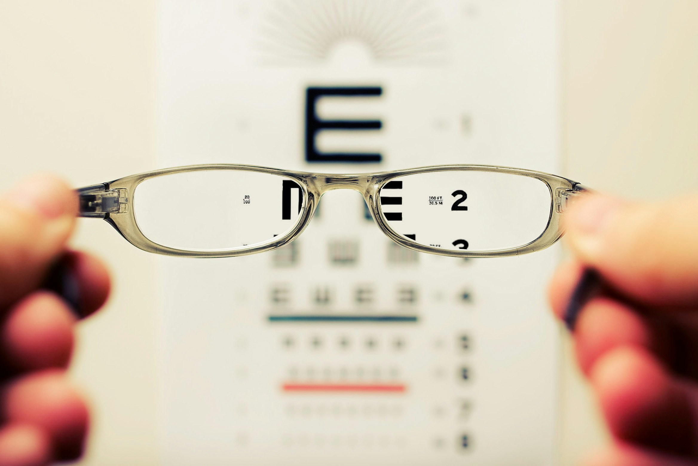 A pair of glasses being held up in front of an eye chart. You can see through the lenses to the shapes and letters clearly. The rest of the chart outside the lenses is blurry.