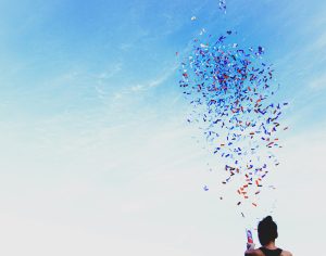 The head and shoulders of a woman facing a blue sky. She is holding a tube from which emerges a cloud of red, white, and blue confetti.