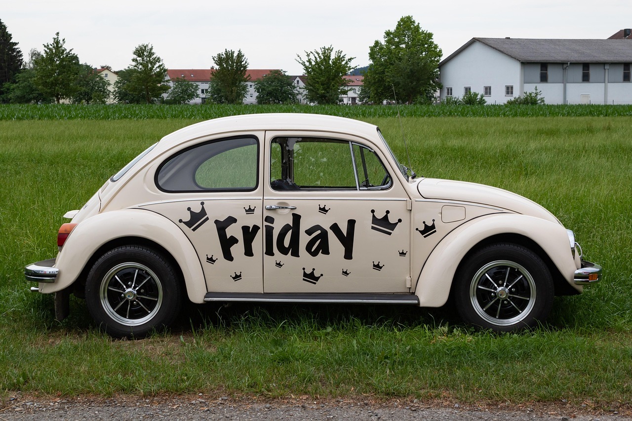A classic VW Beetle, cream in color, with the word 'Friday' painted on the side. It sits in an open grassy field.