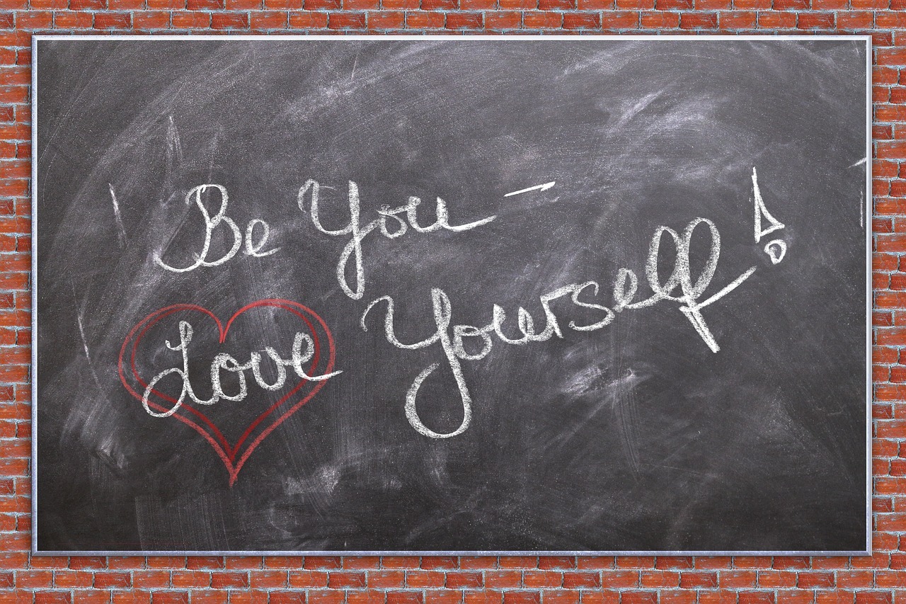 A chalkboard with a brick border. Written on the board in script is the phrase "Be you - Love Yourself". The word "Love" has a red heart drawn around it.