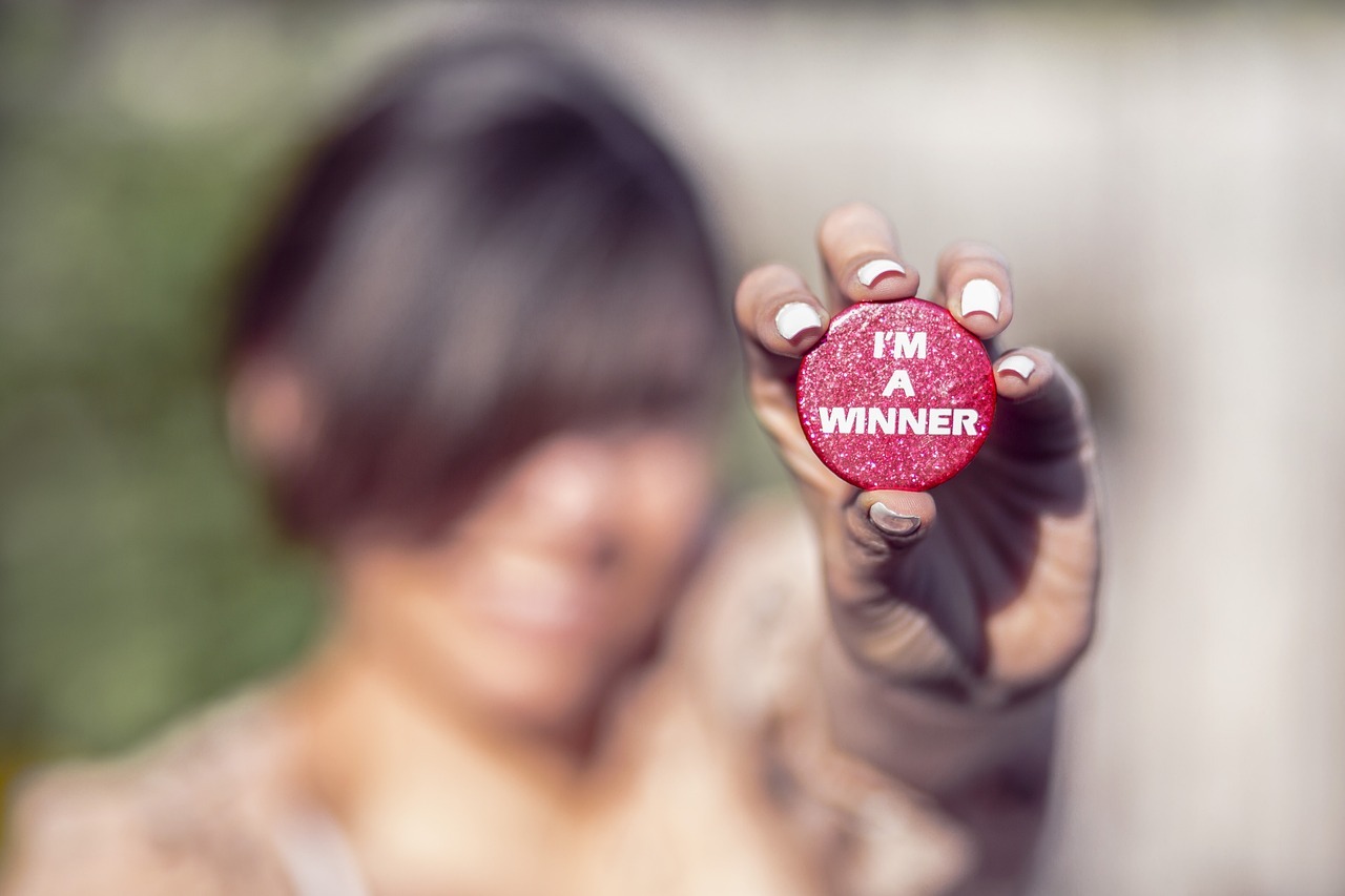A blurry female figure, holding a red wooden disk or chip in her hand out towards the viewer. On the disk are the words "I'm A Winner" in focus