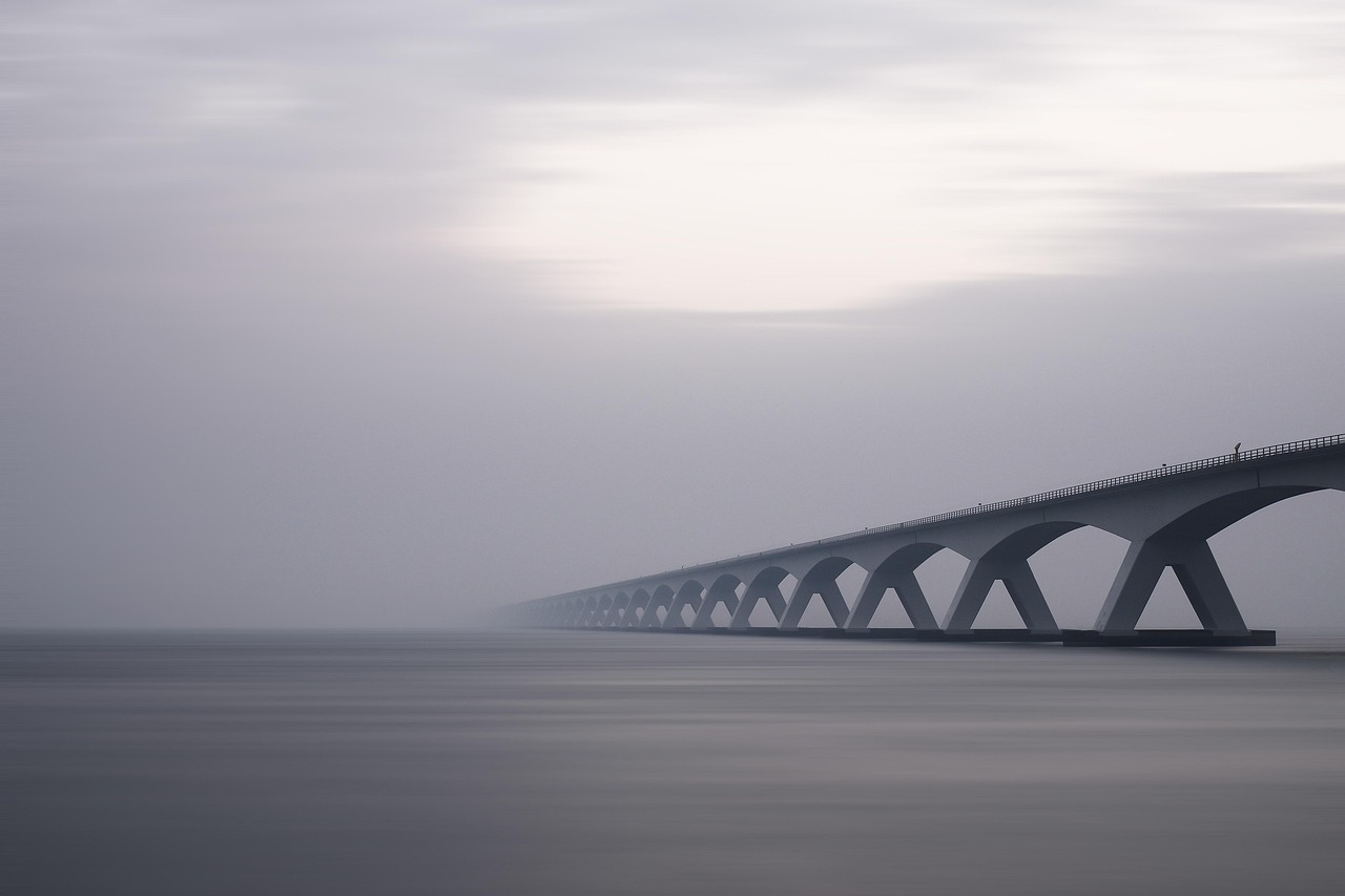 A grey image of a modern-looking arched bridge stretching into the distance, partially shrouded by fog.