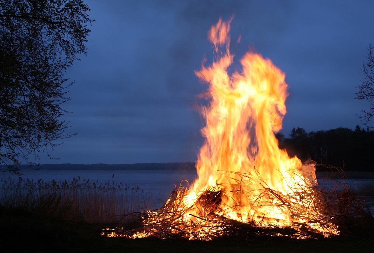 A large bonfire set against an evening sky in front of a lake.