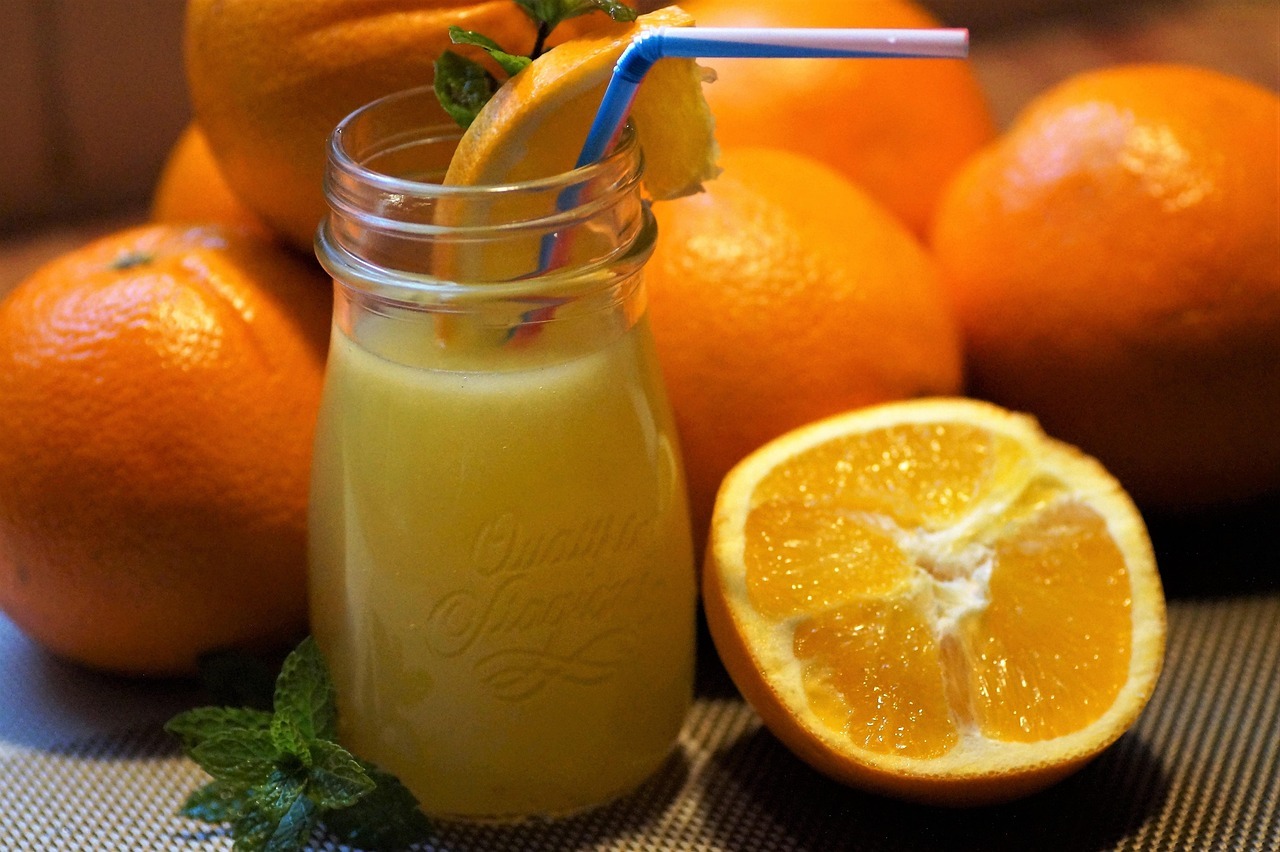 A glass of fresh orange juice with a straw in front of several whole oranges. One orange is sliced in half next to the glass.