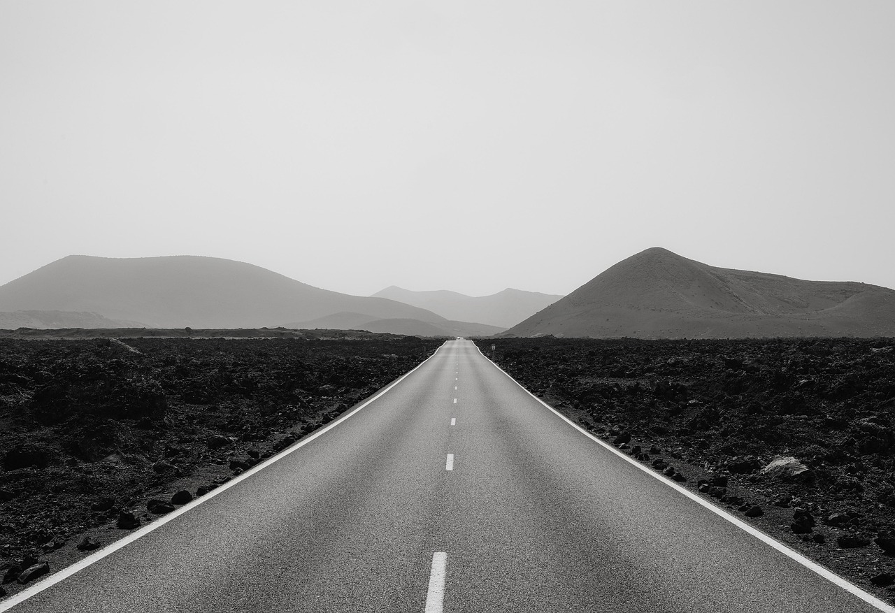 A black and white image of a two-lane road leading off into the horizon. The road bisects hills in the distance.