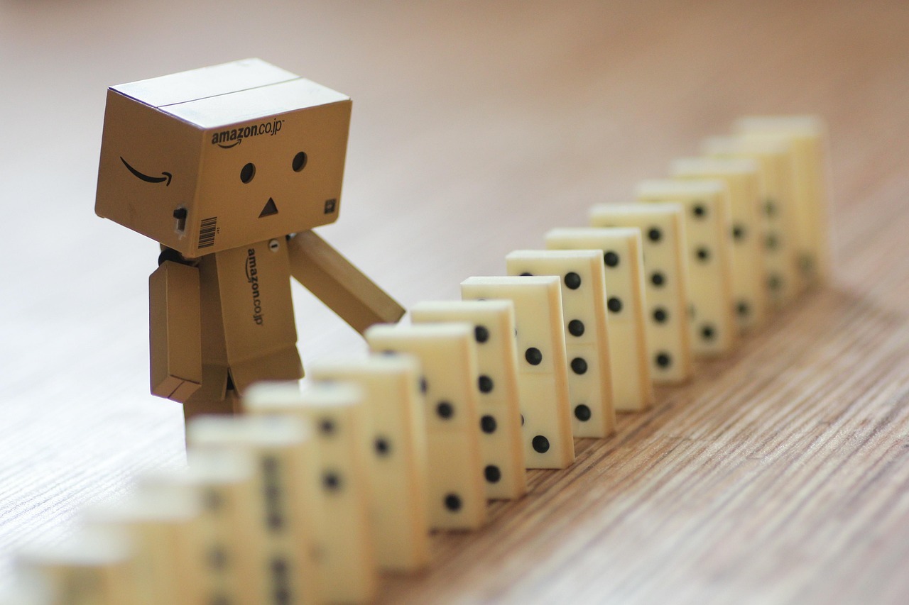 A line of dominos waiting to be jiggled into falling, with a cardboard figure of a box-person reaching out to start the fall.